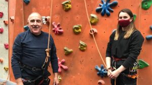 A photo of John stood by a climbing wall, with a rope ready to climb. A female instructor is stood next to him