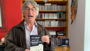 A photo of Steve Woodhull holding his book. He has grey hair and glasses and is wearing shirt and a brown leather jacket. Behind him is a red wall and a bookcase with CDs and DVDs on.