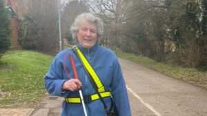 A photo of Margaret. She has a high vis band on is holding a long cane