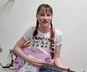 Lily is sat on a stool with a pink electric guitar. She is wearing jeans and a minnie mouse t- shirt. She has her hair in two plaits.