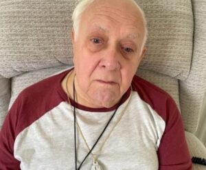 Mike sat in an armchair he is wearing a red and white top and has short white hair.