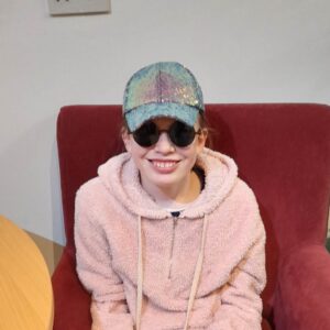 Clara is sat on an armchair. She is wearing a pink hoodie, baseball cap and dark glasses.