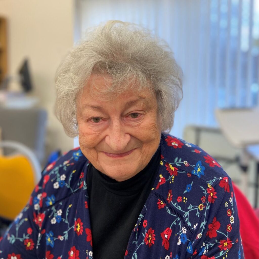 Valerie looks at the camera smiling. She has short grey hair and is wearing a blue floral blouse.