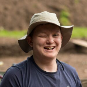 Jonathan- a young man- smiles at the camera. He's wearing a beige bucket hat and a dark blue T-shirt.
