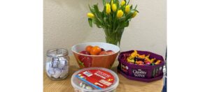 A photo of a small table. ON the table is a vase with yellow tulips in. a bowl of fruit, a box of quality street, a tub of haribo sweets and a jar with folded pieces of paper in