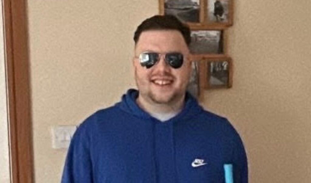 Matthew in a blue top wearing sunglasses and holding a white cane