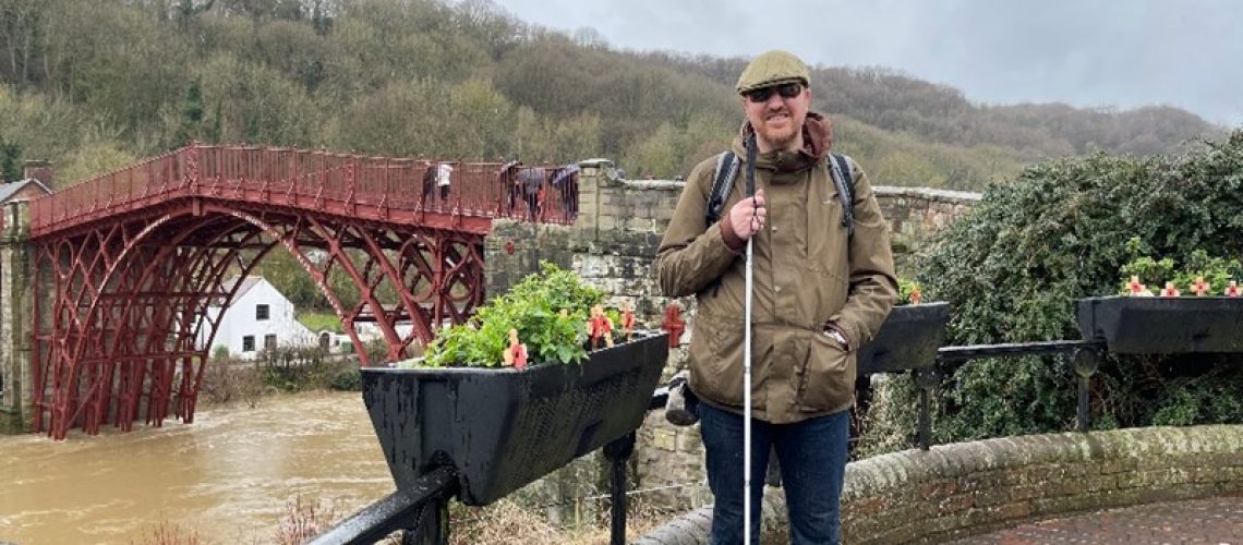Chris is wearing a flat cap, dark glasses, a green coat, jeans and a back pack. He is holding a long white cane. In the background is IronBridge and a wooded hillside. The sky is grey. Next to Chris is a black metal fence with a black flower planter on it with green plants in it and red poppies. Under the bridge a murky river is flowing and a small white cottage is on the river bank.
