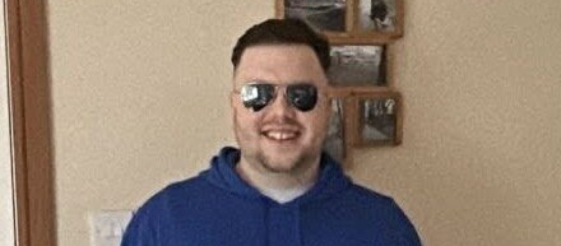 Matt in a blue jumper, wearing glasses and holding his white cane