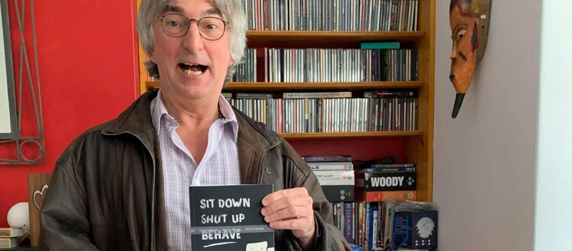A photo of Steve Woodhull holding his book. He has grey hair and glasses and is wearing shirt and a brown leather jacket. Behind him is a red wall and a bookcase with CDs and DVDs on.