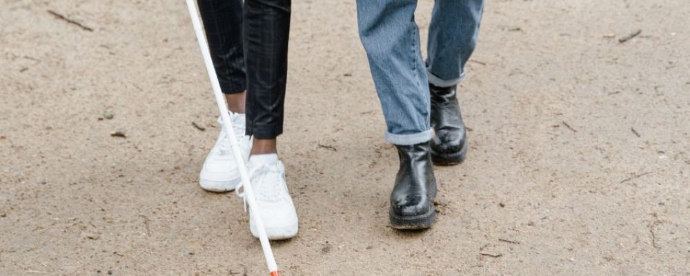 the legs of 2 people walking down the street next to each other. One of the people is holding a white cane.