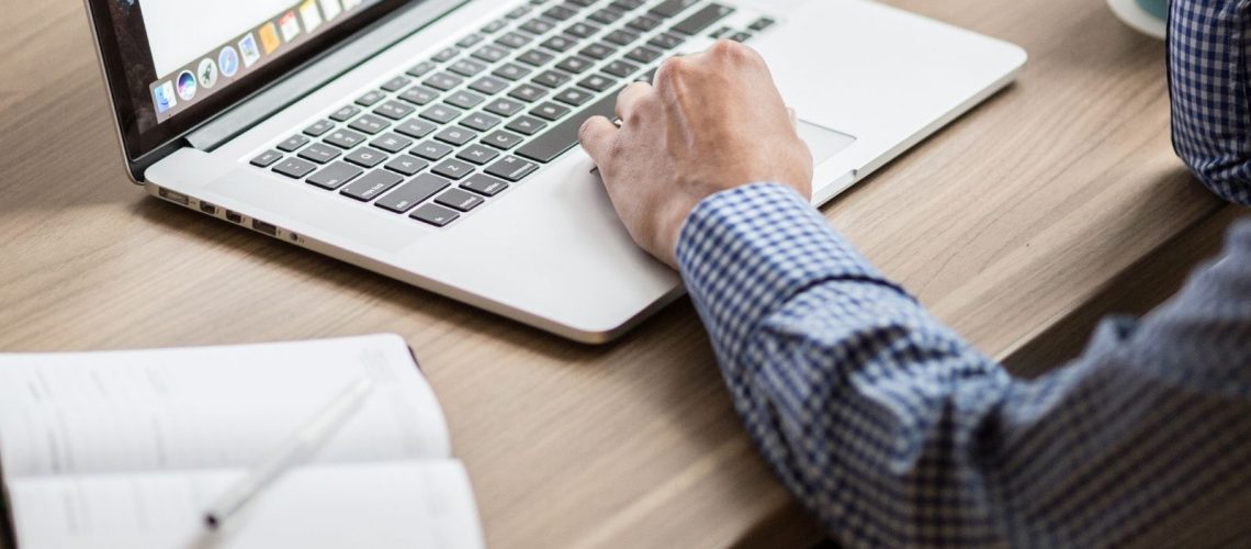 a laptop with a note pad and pen next to it. A man has his hands on the keyboard of the laptop